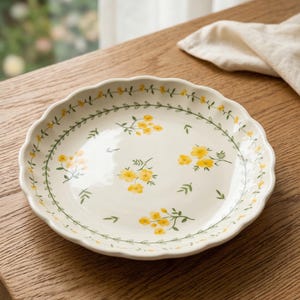 May include: A white ceramic plate with a scalloped edge. The plate is decorated with yellow floral patterns and green leaves. A green vine with leaves and small yellow flowers borders the edge of the plate. The plate is on a wooden surface.