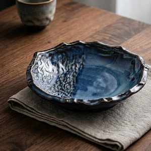 Peut inclure: Un bol en céramique bleu fait à la main avec un motif texturé en forme de vagues sur le bord et l'intérieur. Le bol est posé sur une serviette en lin grise, avec une table en bois en arrière-plan. Une petite tasse en céramique assortie est également visible.