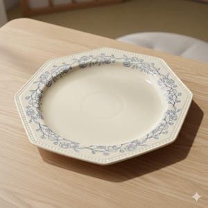 Peut inclure: Une assiette octogonale en céramique blanc cassé avec un motif floral bleu délicat sur le bord. L'assiette a un bord perlé et repose sur une surface en bois clair. Une pièce classique pour servir ou décorer.