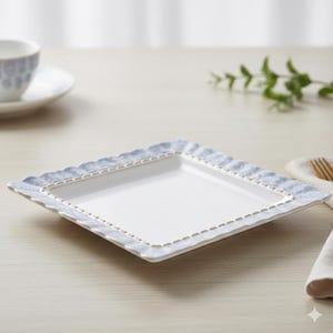 Peut inclure: Une assiette carrée blanche avec une bordure festonnée bleu clair et une bordure dorée pointillée. L'assiette est posée sur une surface en bois de couleur claire. Une tasse et une soucoupe assorties sont visibles en arrière-plan.
