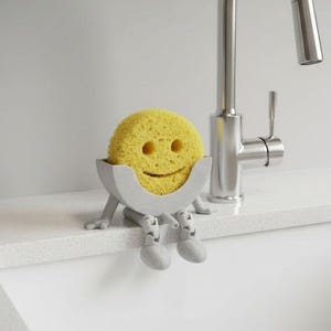 Puede incluir: Una esponja amarilla con una cara sonriente se encuentra en un soporte gris con patas y pies, posada en el borde de un fregadero blanco. Un grifo plateado está en el fondo. El soporte para esponja es un accesorio de cocina divertido y funcional.