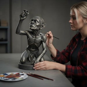 May include: A detailed, metallic sculpture of a kneeling, emaciated figure with large ears, holding a small object aloft. The figure is being painted with a fine brush. A palette of paints and brushes are on the table.