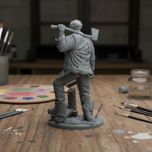 May include: A gray figurine of a figure in a hockey mask holding an axe over his shoulder. The figure is wearing a torn jacket and pants, standing on a small base with wooden posts and cattails. The figurine is on a wooden table with art supplies.