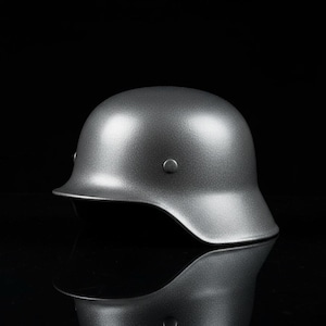 Puede incluir: Un casco gris metálico con una parte superior redondeada y un borde. El casco tiene una superficie lisa y reflectante y está sobre un fondo negro. El diseño del casco incluye un pequeño remache circular en el lateral.