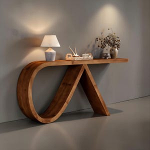 May include: A wooden console table with a unique, curved design. The table features a light-colored lamp, a woven tray with decorative items, and a vase with dried flowers. The table's warm wood tones create a welcoming atmosphere.