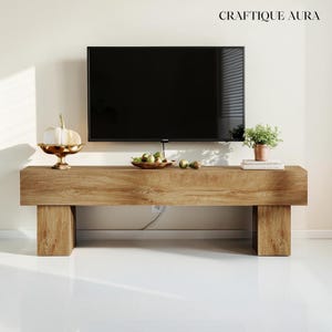 May include: A modern, rectangular wooden TV stand with a natural wood grain finish. The stand features a large, black flat-screen TV mounted above it. Decorative items include a gold-colored pedestal bowl with a white pumpkin, a wooden bowl with fruit, and a small potted plant.