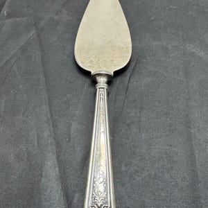 May include: A silver-toned cake server featuring a pointed, triangular blade and a long, tapered handle. The handle is decorated with intricate, embossed floral and geometric patterns, showing a metallic finish with visible signs of age and wear.