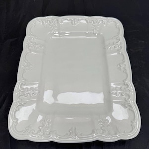 May include: A rectangular white ceramic serving platter featuring a smooth, flat center and a raised, ornate border with intricate, embossed scrollwork and leaf-like patterns along the edges, finished with a glossy, reflective glaze.