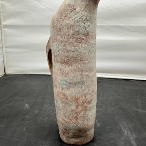 May include: A tall, textured ceramic pitcher with a curved spout and a handle. The pitcher features a repeating wave pattern in shades of brown, beige, and hints of blue. The handmade pottery has a rustic, artisanal aesthetic.