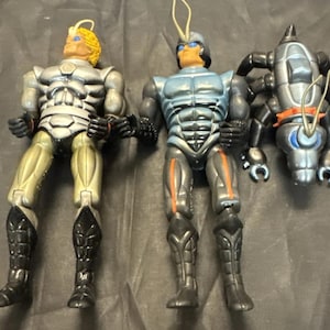 May include: Three action figures are displayed. The figures are humanoid with insect-like features. One figure is silver and gold, another is blue and black, and the third is a smaller, silver and black insectoid robot.