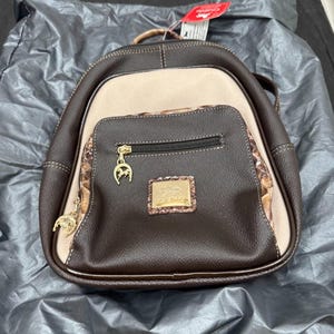 May include: A dark brown and beige backpack featuring a front zippered pocket with a gold-toned horseshoe charm. The bag includes a rectangular gold-toned emblem with an embossed horse design and textured brown trim along the pocket edges.