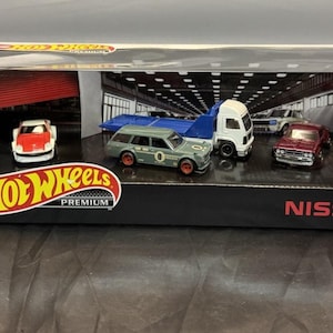 May include: A Hot Wheels Premium Nissan die-cast car set in a display box. The set includes several miniature cars, a truck, and a car carrier. The box features the Hot Wheels logo and the Nissan brand name.