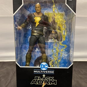 May include: A Black Adam action figure in a clear plastic display case. The figure is wearing a black and gold costume with a lightning bolt emblem. The packaging features the DC Multiverse logo and the words "Black Adam". The age range is 12+.