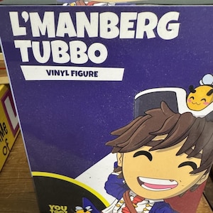May include: A rectangular purple product box for a L'Manberg Tubbo vinyl figure by Youtooz. The box features white text reading L'MANBERG TUBBO VINYL FIGURE, a cartoon illustration of a character with brown hair, and two small yellow bee icons.