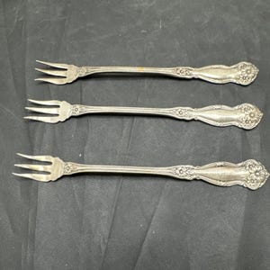 May include: Three silver-toned metal cocktail forks with three tines each. The handles feature an embossed floral pattern at the base and a smooth, tapered midsection. The forks have a metallic finish and a slender, elongated design.