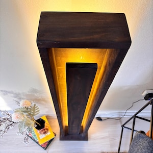May include: A dark brown wooden floor lamp with a rectangular design. The lamp has a warm yellow light emanating from within its geometric structure. The lamp is placed on a light-colored wooden floor.