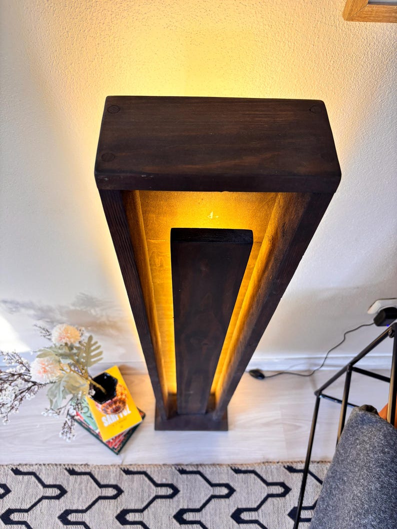 May include: A dark brown wooden floor lamp with a rectangular design. The lamp has a warm yellow light emanating from within its geometric structure. The lamp is placed on a light-colored wooden floor.