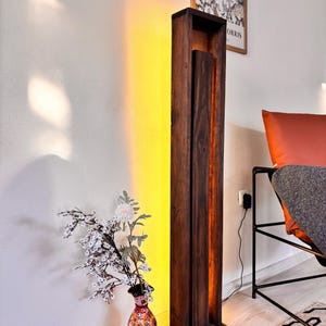 May include: A tall, dark brown wooden floor lamp with a rectangular design. The lamp has a vertical, open-faced structure with a warm, yellow light emanating from within. A vase of flowers sits on a stack of books nearby.