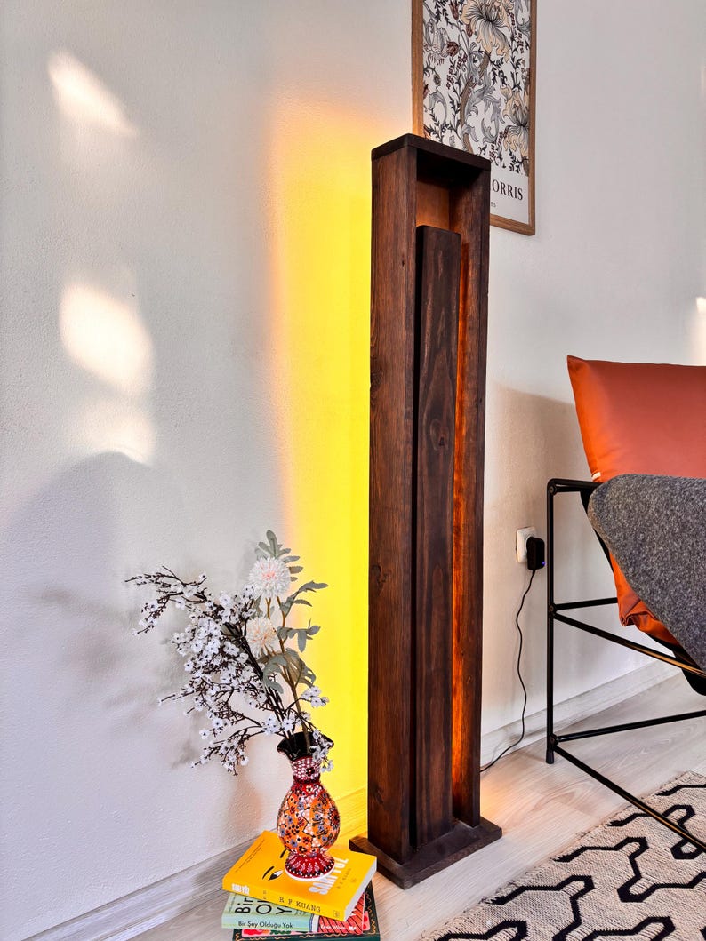 May include: A tall, dark brown wooden floor lamp with a rectangular design. The lamp has a vertical, open-faced structure with a warm, yellow light emanating from within. A vase of flowers sits on a stack of books nearby.