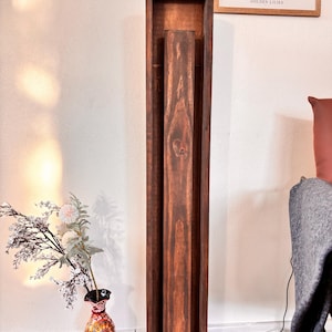 May include: A tall, dark brown wooden floor lamp with a rectangular frame and a central, vertical wooden element. The lamp has a rich, warm tone and stands on a square base. The lamp is next to a vase with flowers and a framed print.