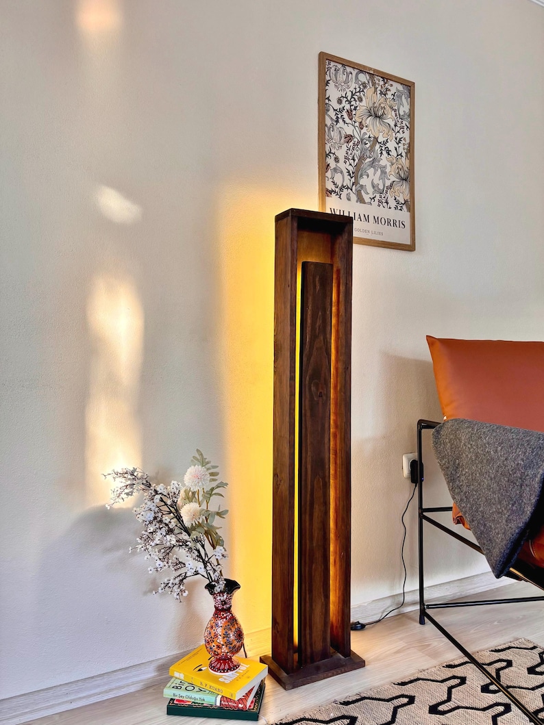 May include: A tall, rectangular wooden floor lamp with a warm, glowing light emanating from the center. The lamp is made of dark wood and stands next to a vase of flowers and a framed print on the wall. The lamp is approximately 60 inches tall.