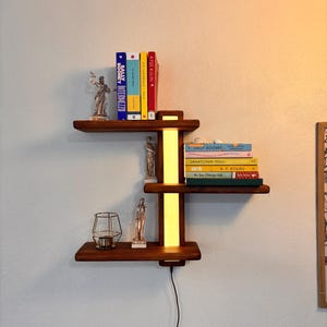 以下が含まれることがあります： 内蔵照明付きの、濃い色の木製3段ウォールシェルフ。本、小さな像、装飾的なキャンドルホルダーが置かれています。シェルフはコンセントに接続されています。