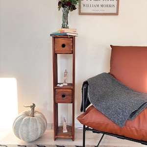 Könnte beinhalten: Ein hoher, schmaler Holzschrank mit zwei Schubladen, in denen kleine Statuen stehen. Auf dem Schrank stehen eine Vase mit Blumen und Bücher. Ein Kürbis und ein gemusterter Teppich befinden sich im Vordergrund. Ein gerahmter William Morris-Druck hängt an der Wand.