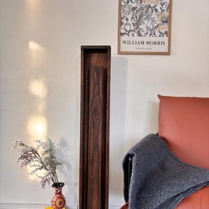 May include: A tall, dark brown wooden floor lamp with a rectangular frame. The lamp is next to a red leather armchair with a gray blanket draped over it. A framed William Morris print hangs on the wall.