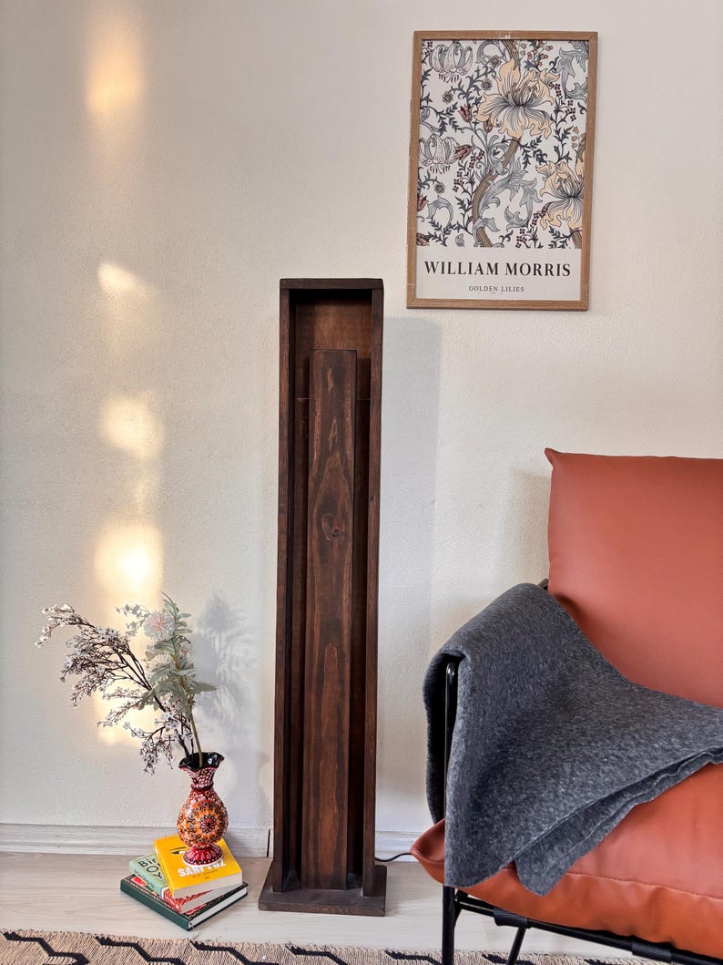May include: A tall, dark brown wooden floor lamp with a rectangular frame. The lamp is next to a red leather armchair with a gray blanket draped over it. A framed William Morris print hangs on the wall.