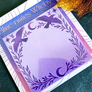 Peut inclure: Une pile de blocs-notes "Bloc-notes Witchy". Le bloc-notes supérieur présente un fond dégradé violet avec un motif de corbeau et de croissant de lune. Le bloc-notes est bordé d'un motif de vigne feuillue. Le bloc-notes inférieur a une bordure rayée bleue et blanche.