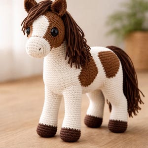May include: A handmade crochet horse toy in white and brown. The horse has a white body with brown spots, a brown mane and tail, and brown hooves. The toy is standing on a wooden surface.