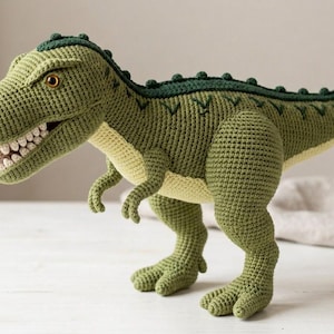 May include: A crocheted Tyrannosaurus Rex dinosaur toy featuring a sage green body with a cream-coloured underbelly. The dinosaur has a dark green spine with small bumps, a wide open mouth showing white teeth, and amber-coloured eyes.