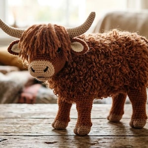 Puede incluir: Figura de vaca de las Tierras Altas hecha a mano con ganchillo. La vaca marrón tiene un pelaje lanudo, cuernos largos y un flequillo esponjoso. Está sobre una superficie de madera. Es un objeto decorativo encantador.