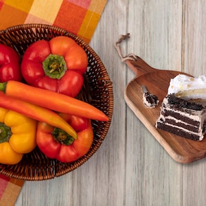 May include: A wicker basket containing red and yellow bell peppers and orange chilli peppers. Beside the basket is a wooden chopping board with two slices of cake and a small spoon.