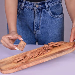 May include: A wooden serving board with a rectangular shape, holding several small, brown, rectangular crackers. One cracker is being held by a person. The board has a natural wood grain and a rope handle.