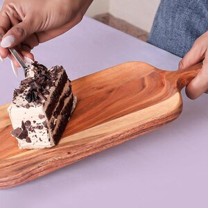 Könnte beinhalten: Ein Stück Schokoladenkuchen mit Cremefrosting und Schokoladenraspeln auf einem Servierbrett aus Holz. Das Brett ist rechteckig mit einem Griff und besteht aus hellem und dunklem Holz.