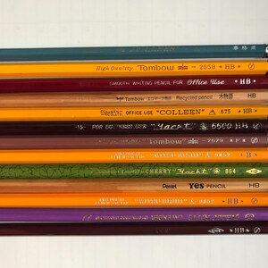 Puede incluir: Una colección de lápices de varios colores y diseños, dispuestos en filas paralelas. Los lápices muestran varias marcas y textos, incluyendo "Tombow" y "Yes PENCIL". Cada lápiz tiene una goma de borrar y una banda metálica.