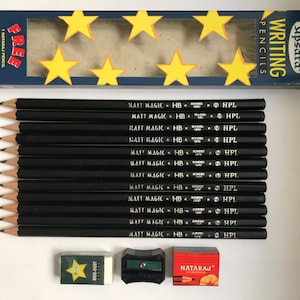 Puede incluir: Un conjunto de lápices negros con la inscripción "MATT MAGIC HB HPL", junto con un sacapuntas, una goma de borrar sin polvo y una caja de lápices con recortes de estrellas. La caja dice "WRITING PENCILS" y "FREE".