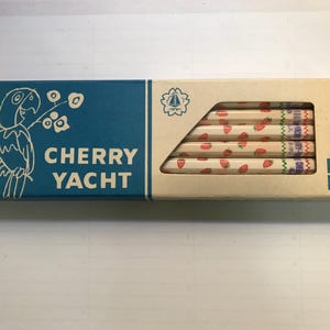以下が含まれることがあります： 「CHERRY YACHT」の文字とオウムのイラストが入った、ヴィンテージの鉛筆箱。鉛筆はイチゴ模様で装飾されています。箱は青とベージュです。