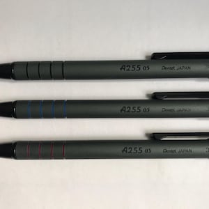 Puede incluir: Tres portaminas grises oscuros con puntas y clips negros. Cada lápiz tiene el texto "A255 05" y "Pentel JAPAN" impreso en el cuerpo. Un lápiz tiene anillos azules, otro rojos y otro no tiene anillos.