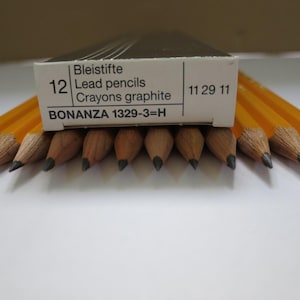May include: A box of 12 yellow hexagonal lead pencils labeled Bonanza 1329-3=H. The box is white with black text, and the pencils are arranged in a row with their sharpened graphite points facing forward, showing the wood grain and lead tips.