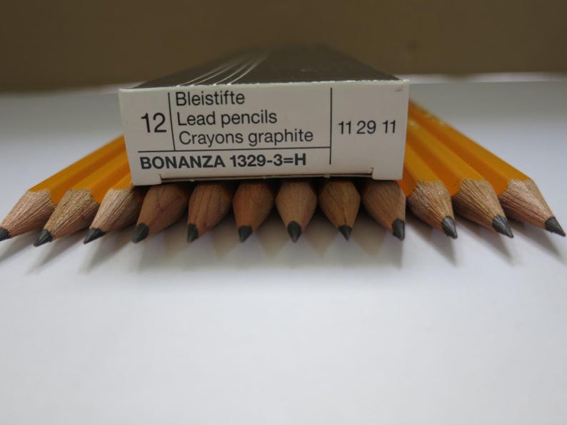 May include: A box of 12 yellow hexagonal lead pencils labeled Bonanza 1329-3=H. The box is white with black text, and the pencils are arranged in a row with their sharpened graphite points facing forward, showing the wood grain and lead tips.