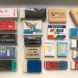 May include: A collection of twenty rectangular and triangular erasers in various colours and brands, including Staedtler, Faber-Castell, Lyra, and Helix. The erasers feature printed text, logos, and graphic illustrations, arranged in a grid on a white surface.