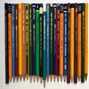 May include: A collection of twenty-five assorted graphite pencils arranged side-by-side in a row. The pencils feature various brand names, including Lyra, Staedtler, and Faber-Castell, with diverse barrel colours such as yellow, orange, blue, green, and black.