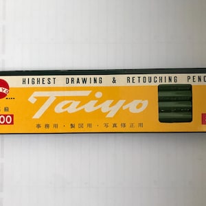 Puede incluir: Una caja de lápices Taiyo 8000. La caja es amarilla con texto negro y rojo, incluyendo "HIGHEST DRAWING & RETOUCHING PENCILS" y "Taiyo". Los lápices son verdes y la caja está marcada con "3B".