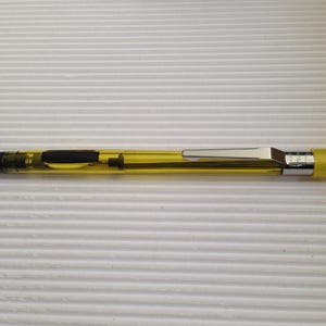Puede incluir: Un portaminas con cuerpo amarillo translúcido, zona de agarre negra y clip de bolsillo de metal plateado. El lápiz presenta una punta cónica negra y una tapa amarilla en el extremo, con componentes internos visibles a través de la carcasa.