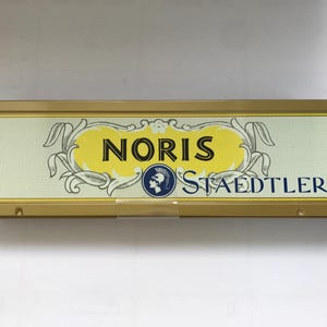 Può includere: Un astuccio rettangolare dorato con le parole "NORIS STAEDTLER" in nero e blu. L'astuccio presenta un design giallo e bianco al centro con il nome del marchio e un logo.