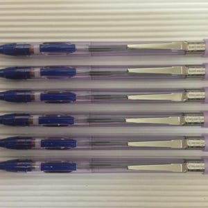 以下が含まれることがあります： 青いインクが入った透明なプラスチック製のペンが5本、上から見た写真。ペンには青いアクセント、シルバーのクリップ、薄紫色の消しゴムが付いています。白い背景に平行に並んでいます。各ペンの長さは約14cmです。