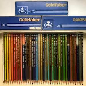 K&ouml;nnte beinhalten: Eine Sammlung von Faber-Castell Goldfaber Bleistiften in einer Pappschachtel, mit drei blauen Schachteln der gleichen Marke dar&uuml;ber gestapelt. Die Bleistifte sind in Regenbogenfarben angeordnet, mit dem Markennamen auf den Schachteln.