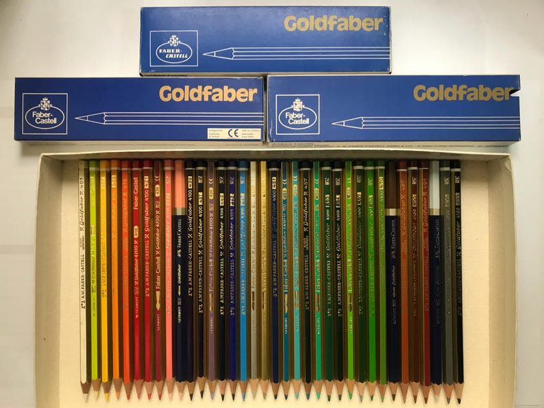 K&ouml;nnte beinhalten: Eine Sammlung von Faber-Castell Goldfaber Bleistiften in einer Pappschachtel, mit drei blauen Schachteln der gleichen Marke dar&uuml;ber gestapelt. Die Bleistifte sind in Regenbogenfarben angeordnet, mit dem Markennamen auf den Schachteln.